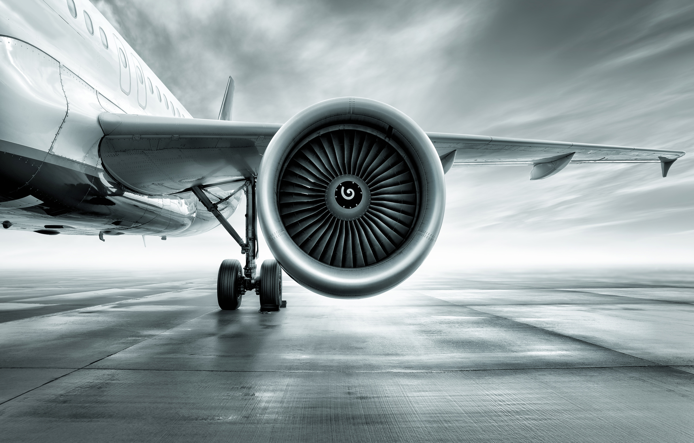 Ein Flugzeugtriebwerk aus der Nähe, das am Boden steht. Die Turbine hat eine spiralförmige Öffnung und ist von einem silbernen Gehäuse umgeben. Im Hintergrund ist das Flugzeugflügel sichtbar, mit einem weiten Himmel über einer glatten Landebahn.