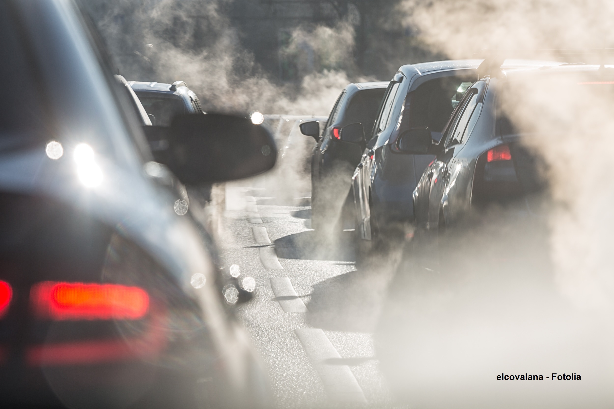 Eine Reihe von parkenden Autos auf einer Straße, umgeben von aufsteigendem Dampf oder Abgasen. Die Szene vermittelt ein Gefühl von Stau und Luftverschmutzung.