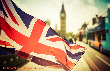 Die britische Flagge weht im Vordergrund, mit unscharfem Hintergrund, der Big Ben und typische Doppeldeckerbusse zeigt. Die Szene vermittelt ein Gefühl von London und britischer Kultur.