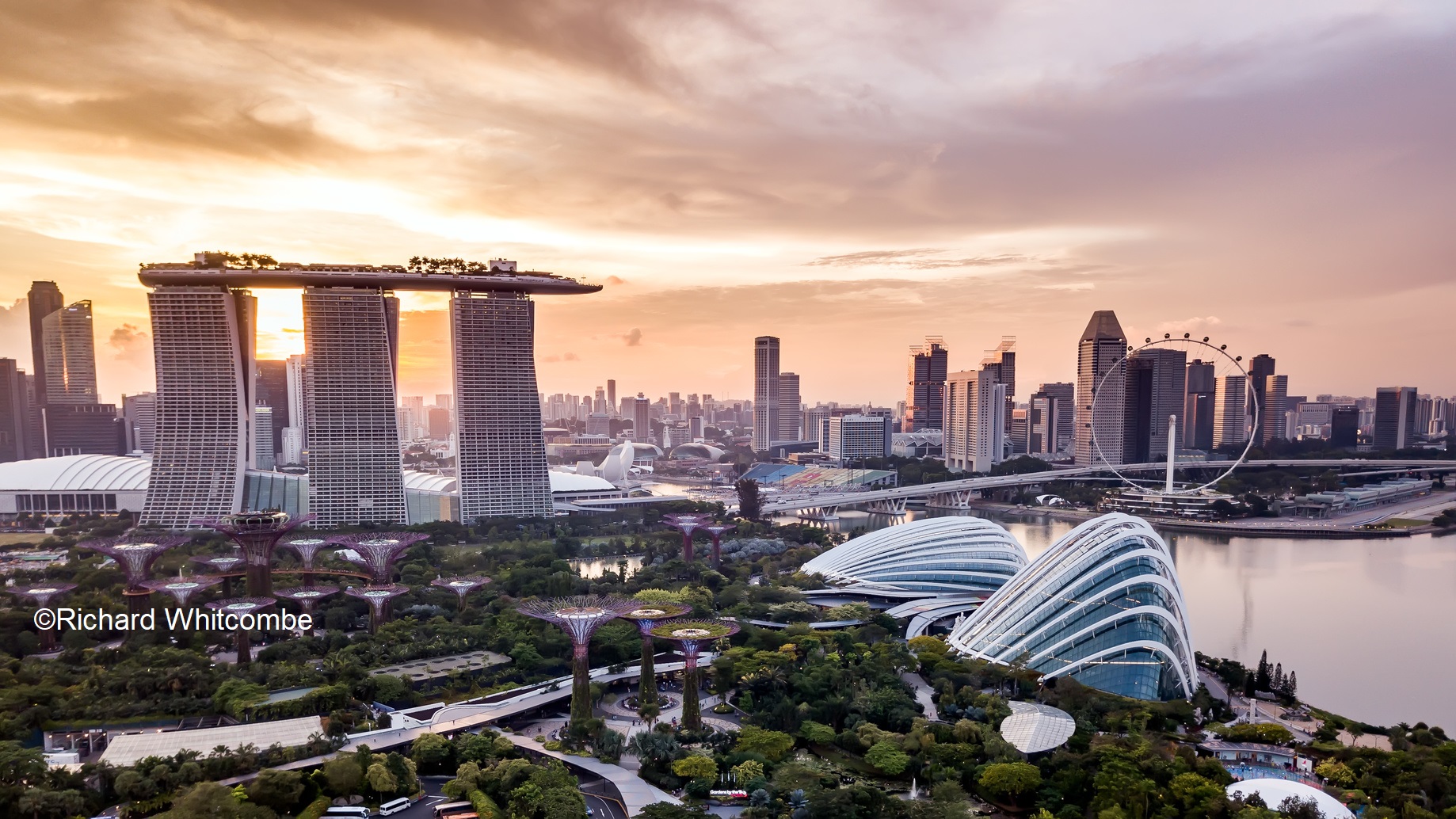 Die Aufnahme zeigt die Skyline von Singapur bei Sonnenuntergang. Im Vordergrund sind futuristische Gebäude, darunter das Marina Bay Sands, umgeben von üppiger Vegetation. Im Hintergrund sind andere Wolkenkratzer und der Singapore Flyer zu sehen, während der Himmel in warmen Farbtönen leuchtet.