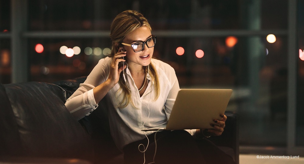 Eine blonde Frau trägt eine Brille und spricht telefonisch, während sie an einem Laptop sitzt. Sie hat Kopfhörer im Ohr und wirkt konzentriert. Die Umgebung ist dunkel, mit unscharfen Lichtern im Hintergrund.