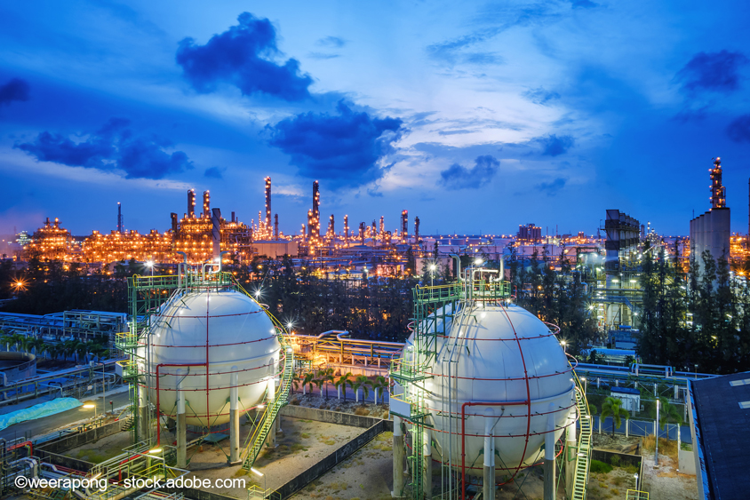 The image showcases a large industrial complex at dusk, featuring two prominent spherical storage tanks in the foreground. The background is lit with numerous illuminated structures and smokestacks, reflecting an active oil or gas refinery amidst a moody blue sky with scattered clouds.