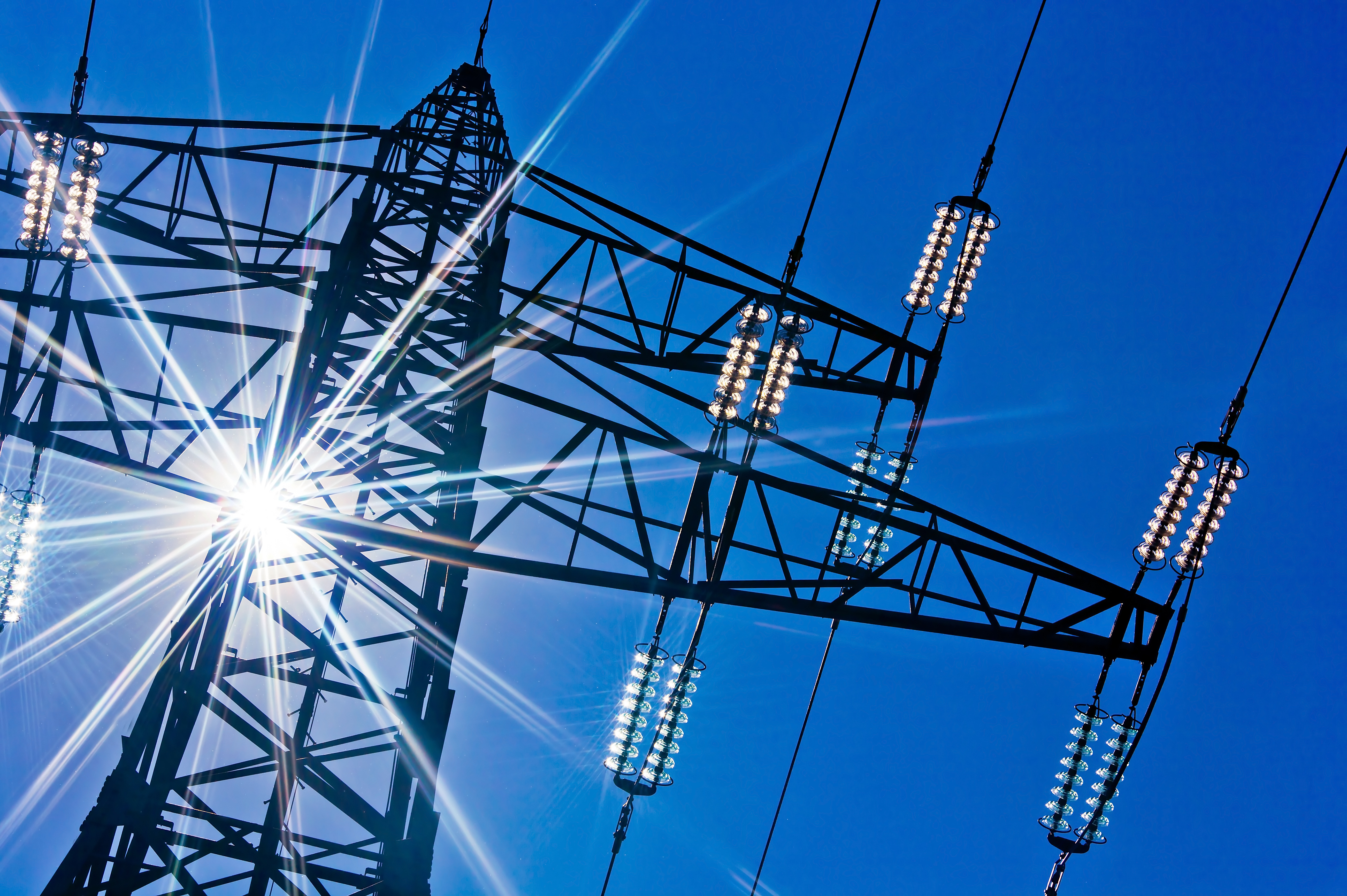 A high-voltage power transmission tower silhouetted against a bright blue sky. Sunlight creates a dazzling flare effect around the tower, enhancing its structure and the surrounding power lines.
