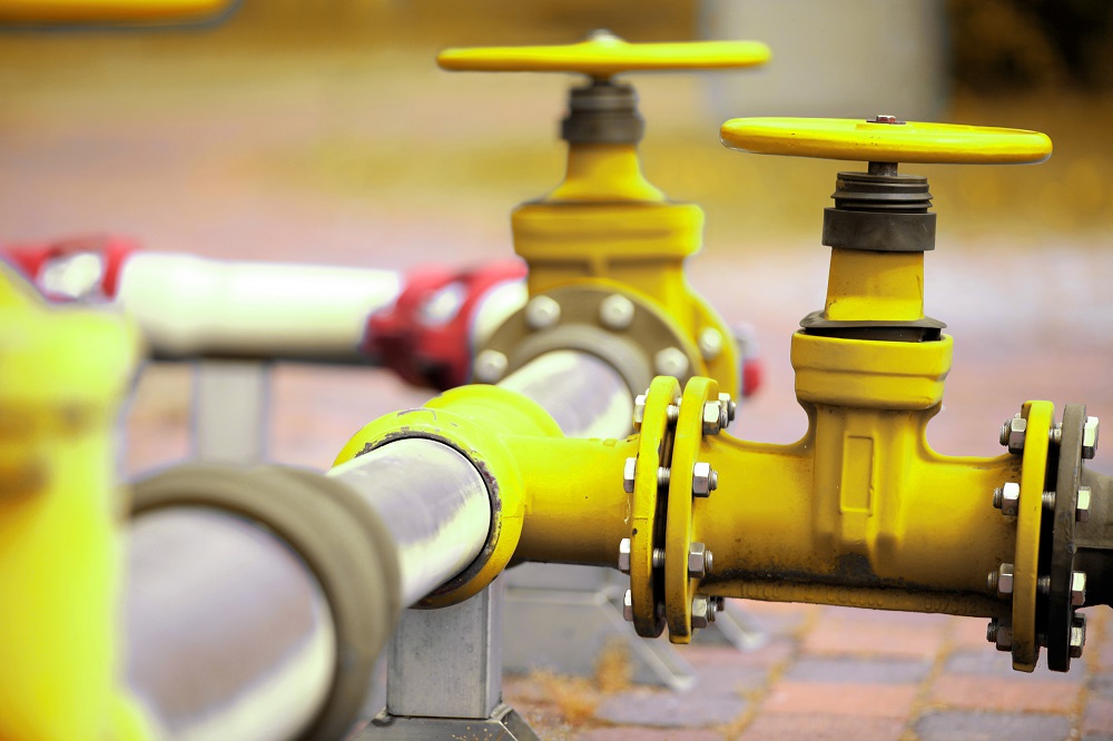 Close-up of a yellow industrial valve and pipeline, showcasing the valve's handle and connection points. The pipes are situated on a textured surface, emphasizing their functional design in a utility setting.