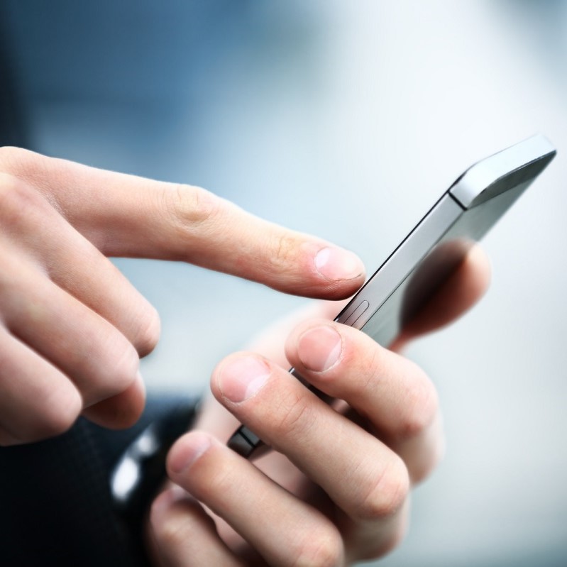 A close-up of hands holding a smartphone. One hand is pressing the screen while the other supports the device. The focus is on the interaction with the phone, suggesting communication or browsing. The background is blurred, emphasizing the activity.