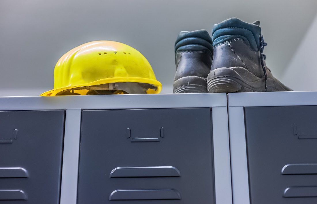 Auf einem grauen Spind liegen ein gelber Sicherheitshelm und ein Paar Arbeitsschuhe. Der Helm ist rund und leuchtend, während die Schuhe robust und abgenutzt wirken. Die Szene vermittelt ein Gefühl von Arbeitssicherheit und Berufstätigkeit.