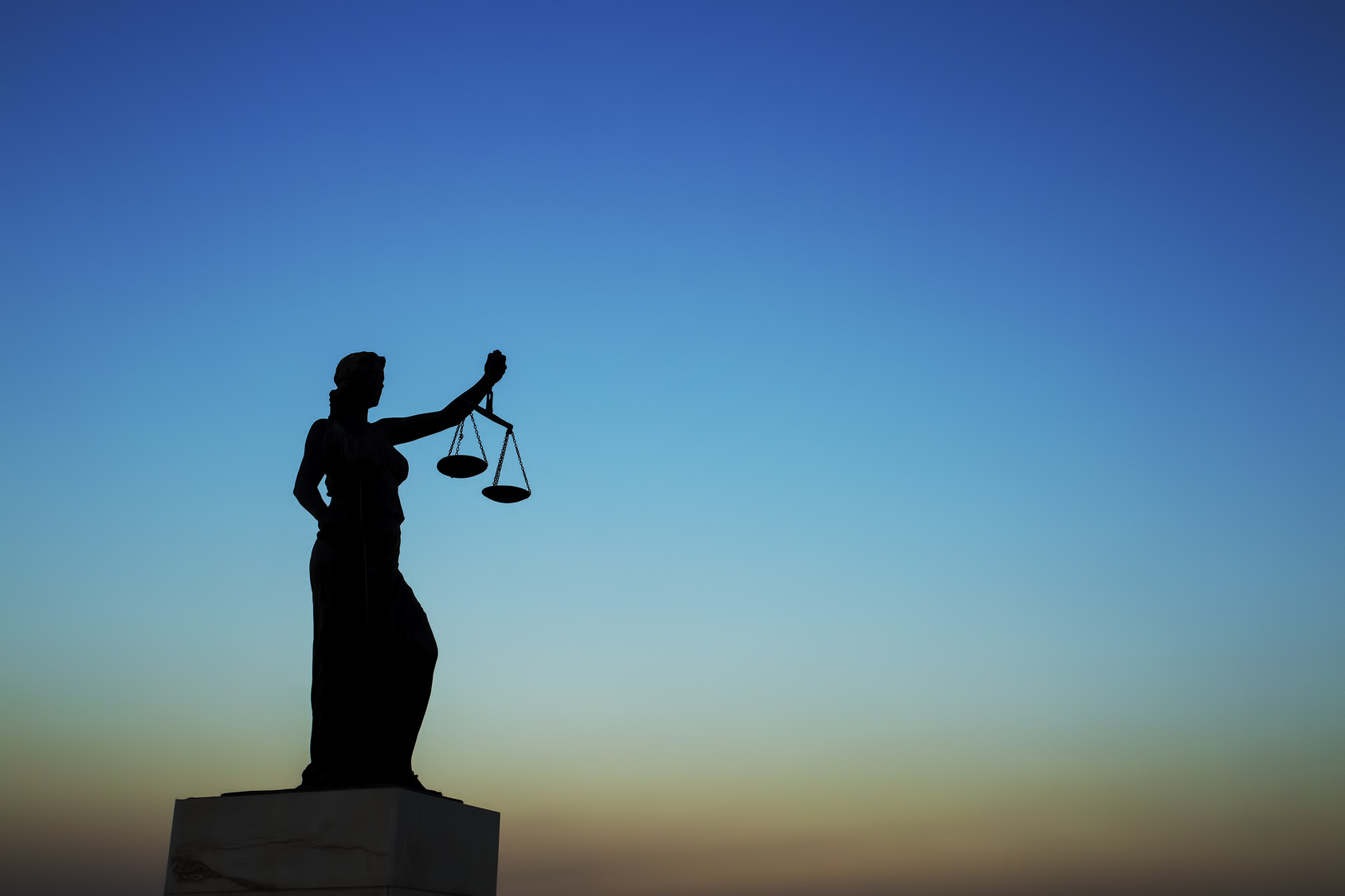 Eine silhouette einer Justitia-Statue, die eine Waage in der Hand hält. Der Hintergrund zeigt einen Farbverlauf von Blau zu Orange, der den Himmel bei Sonnenuntergang darstellt. Die Statue symbolisiert Gerechtigkeit und Fairness.