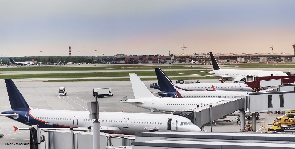 Auf dem Bild sind mehrere Flugzeuge auf einem Flughafengelände zu sehen, darunter einige am Terminal angekoppelt. Im Hintergrund sind weitere Flugzeuge und Flughafeninfrastruktur sichtbar, während der Himmel einen leicht bewölkten Eindruck macht.