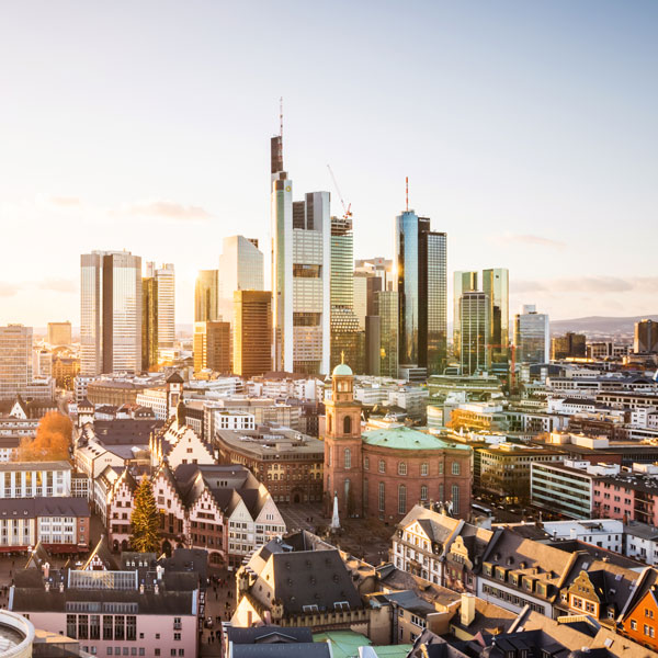 A panoramic view of a city skyline at sunset, featuring modern skyscrapers rising above historical buildings. In the foreground, traditional architecture contrasts with the glass structures, showcasing a blend of old and new. Soft golden light bathes the scene, enhancing its architectural diversity.