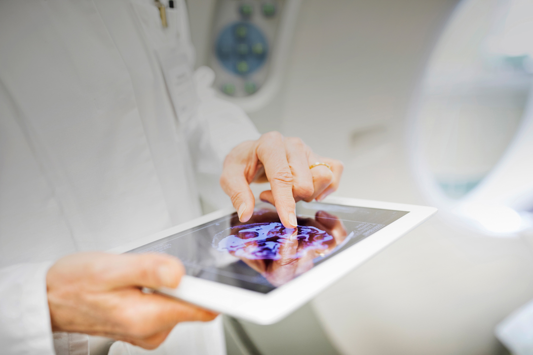 A medical professional in a white coat is holding a tablet displaying an image of a brain scan. Their finger is pointing at the scan, indicating a focus on its details. The background suggests a clinical or healthcare environment.