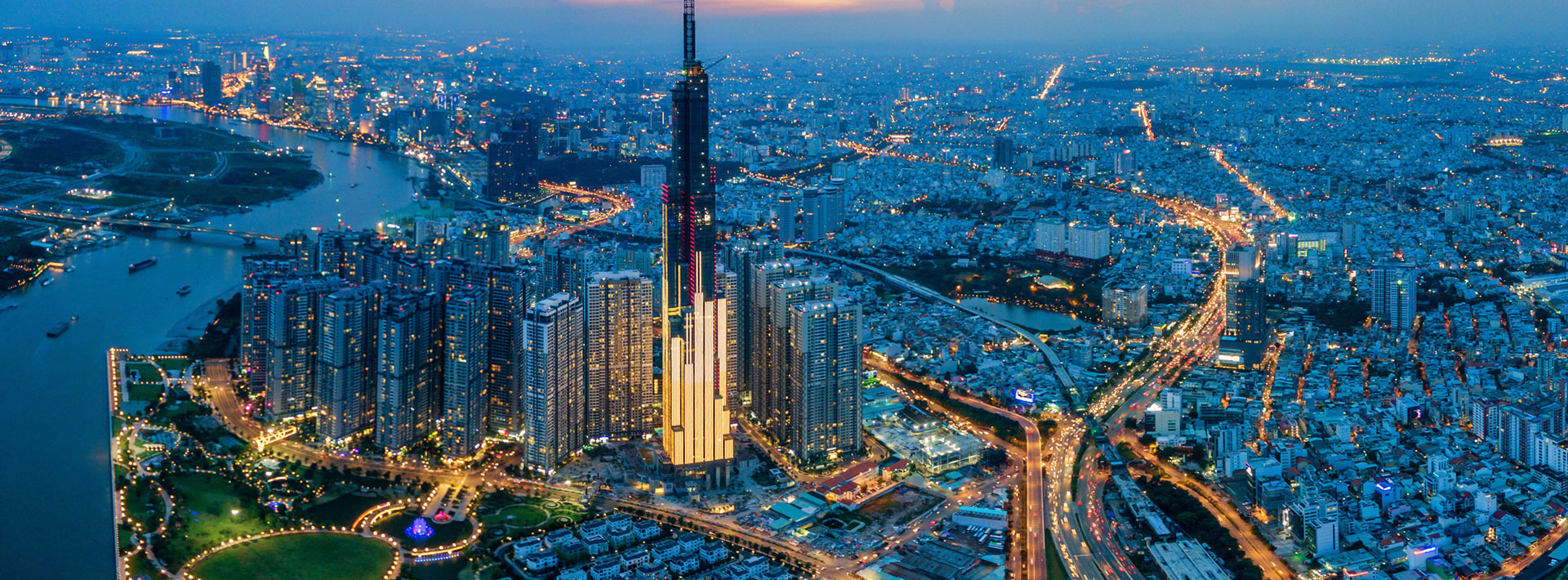 Eine weitläufige Stadtansicht bei Dämmerung, dominiert von einem hohen, beleuchteten Wolkenkratzer. Rund um den Turm erstrecken sich moderne Gebäude und Straßen, während Flüsse und Parkanlagen in der Szene sichtbar sind. Die Stadt glüht mit Lichtern und vermittelt ein Gefühl von Dynamik und urbanem Leben.
