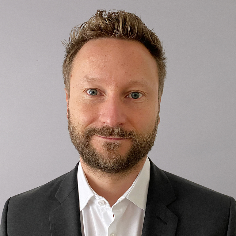 A professional man with light brown hair and a beard is smiling at the camera. He is wearing a black suit jacket over a white shirt. The background is a plain light gray, emphasizing his facial expression and attire.
