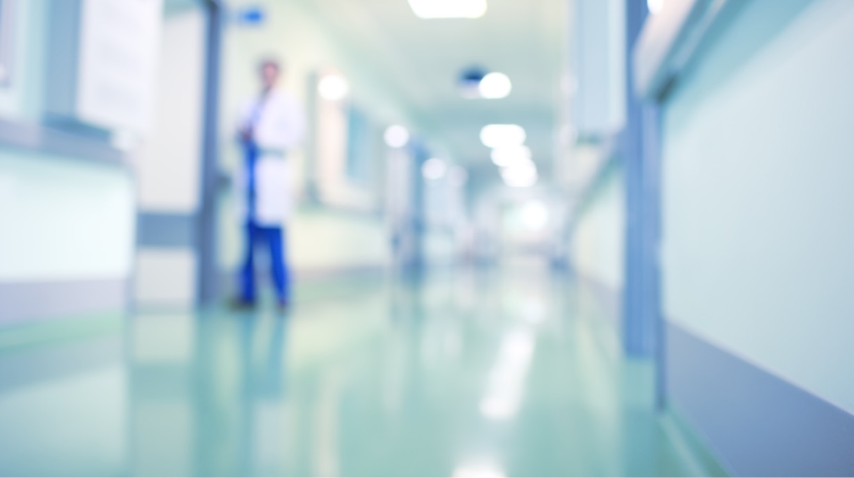 Unscharfe Ansicht eines Krankenhausflurs mit einem Arzt im Hintergrund. Der Flur ist hell erleuchtet und hat grüne Wände sowie einen glänzenden Boden. Der Arzt trägt einen weißen Kittel und steht mit einem verschwommenen Blick im Bild.