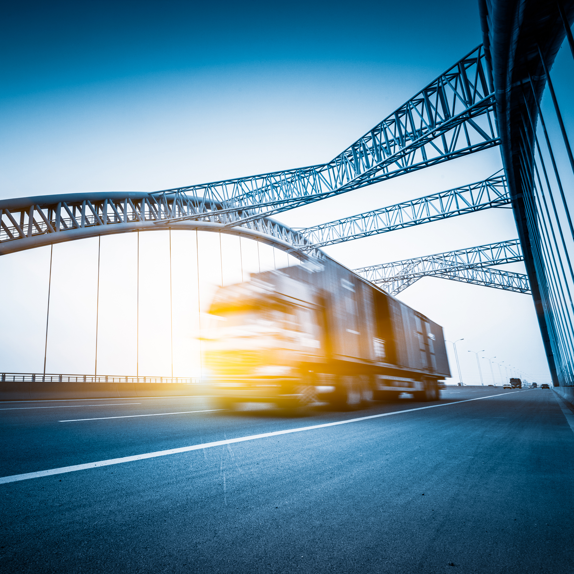Ein Lkw fährt schnell über eine moderne Brücke. Die Struktur der Brücke ist sichtbar, mit geschwungenen Stahlträgern. Der Himmel ist blau mit einem sanften Lichtschein, der die Szene beleuchtet.