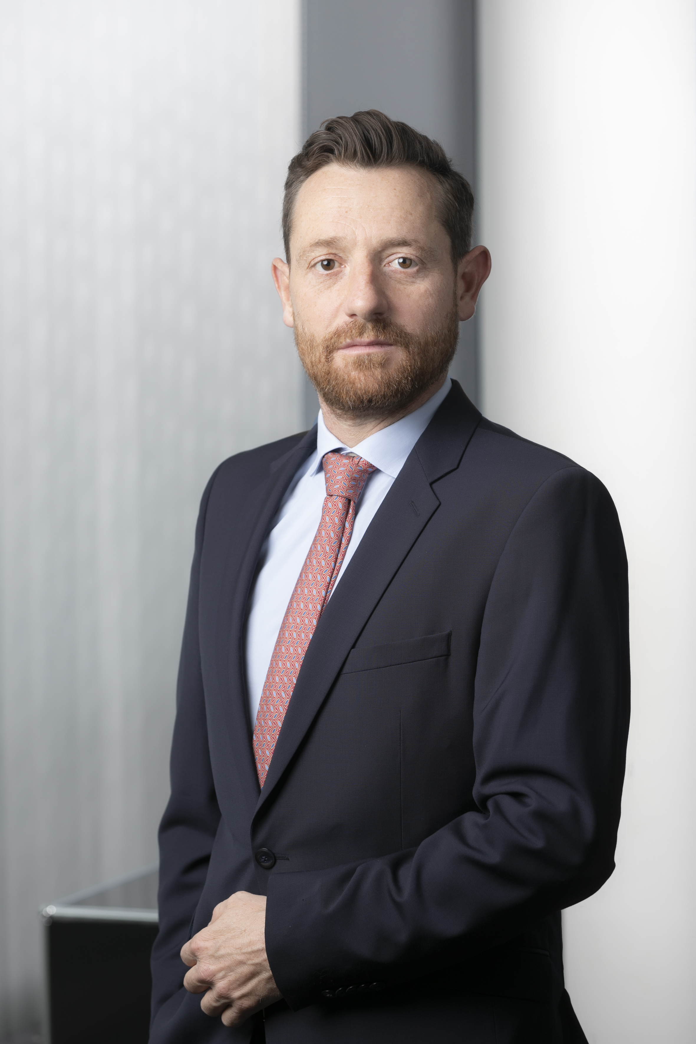 A professional man stands confidently, wearing a dark suit with a light blue shirt and a patterned tie. His expression is serious but approachable, and he stands in front of a softly blurred, light-colored background.