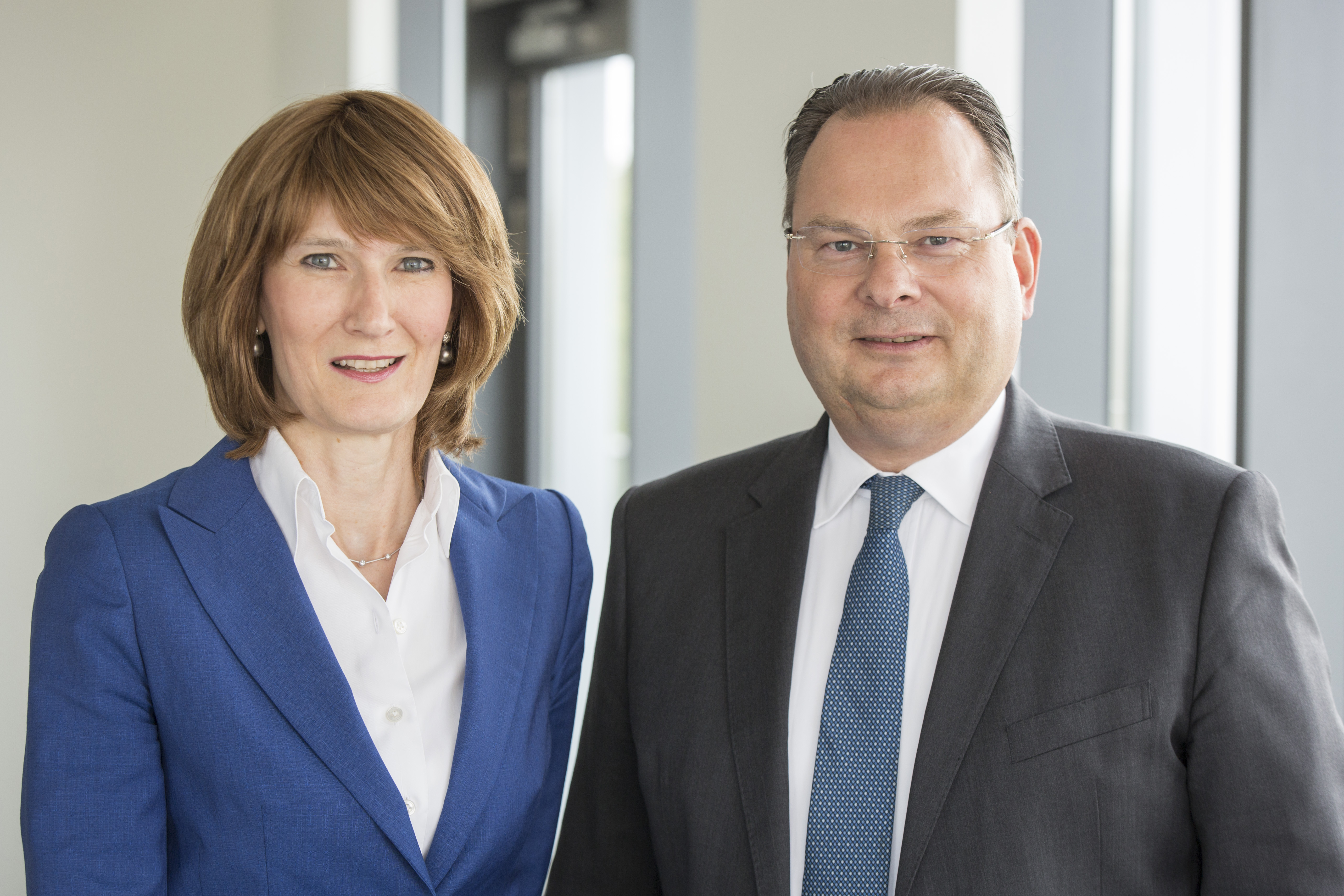 Two professionals stand side by side, both smiling. The woman on the left wears a blue blazer over a white blouse, while the man on the right is dressed in a dark suit with a light-colored shirt and a patterned tie. They appear confident and engaged in a corporate environment.