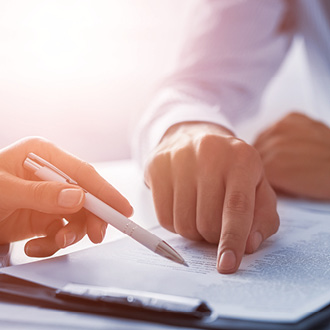 Zwei Hände sind zu sehen, eine hält einen Stift und zeigt auf einen Text auf einem Blatt Papier. Die Szene wirkt professionell und konzentriert, mit sanftem Licht im Hintergrund.