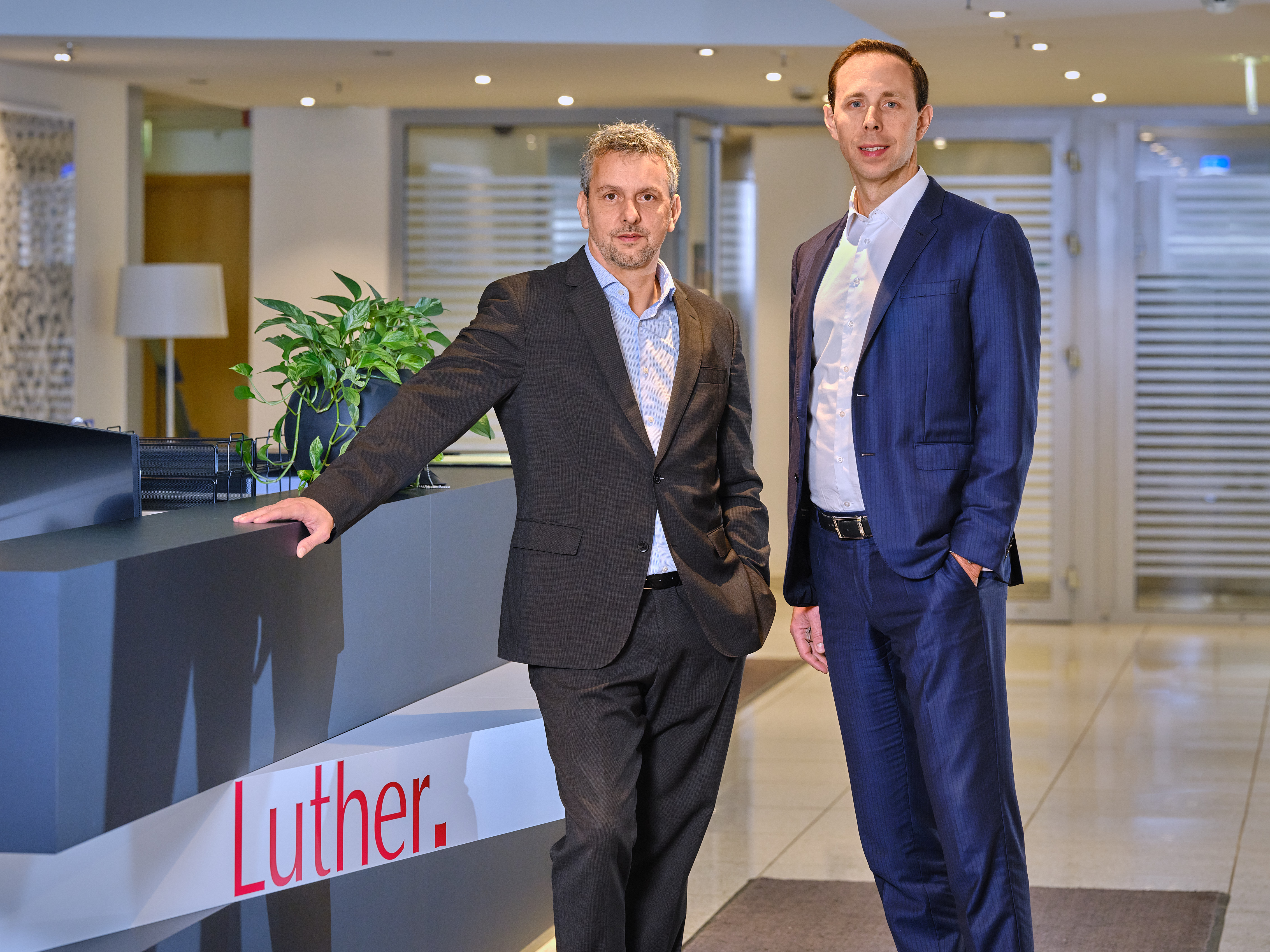 Two professional men stand beside a reception desk in a modern office setting. One man is in a dark suit with a gray shirt, leaning on the desk, while the other is in a blue suit with a white shirt. The desk features the name "Luther." prominently displayed.