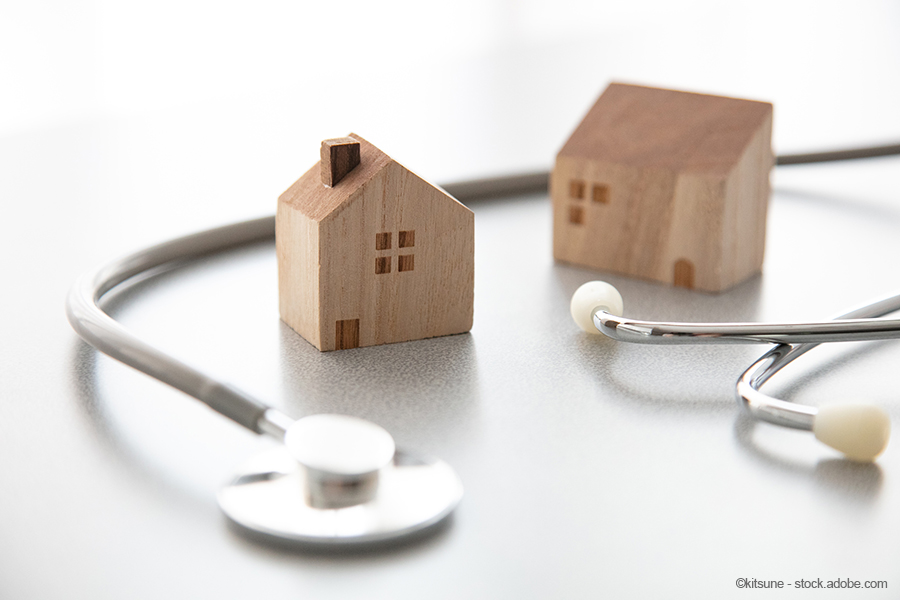 Zwei kleine Holzhäuser stehen neben einem Stethoskop auf einem Tisch. Die Szene vermittelt eine Verbindung zwischen Gesundheit und Heimat, möglicherweise im Kontext der häuslichen Pflege oder Gesundheitsversorgung.