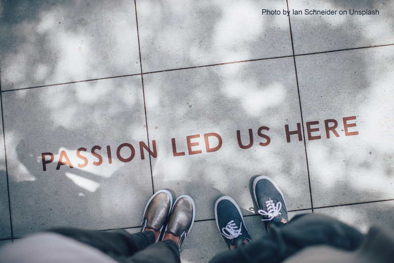 The image features a pair of feet standing on a pavement with the phrase “PASSION LED US HERE” engraved. The text is centrally positioned, surrounded by natural light and shadow from above. The shoes are casual, one pair metallic and the other dark blue.