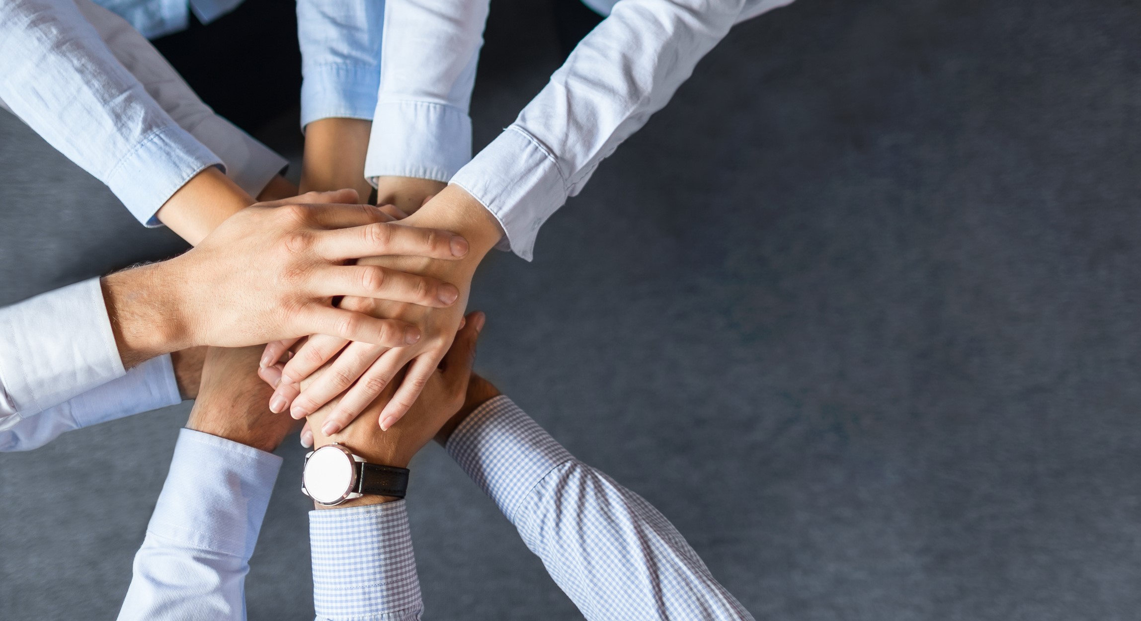 A group of hands from different people is layered on top of each other, symbolizing teamwork and collaboration. The hands are adorned in formal attire and are positioned on a flat surface, conveying unity and collective effort.