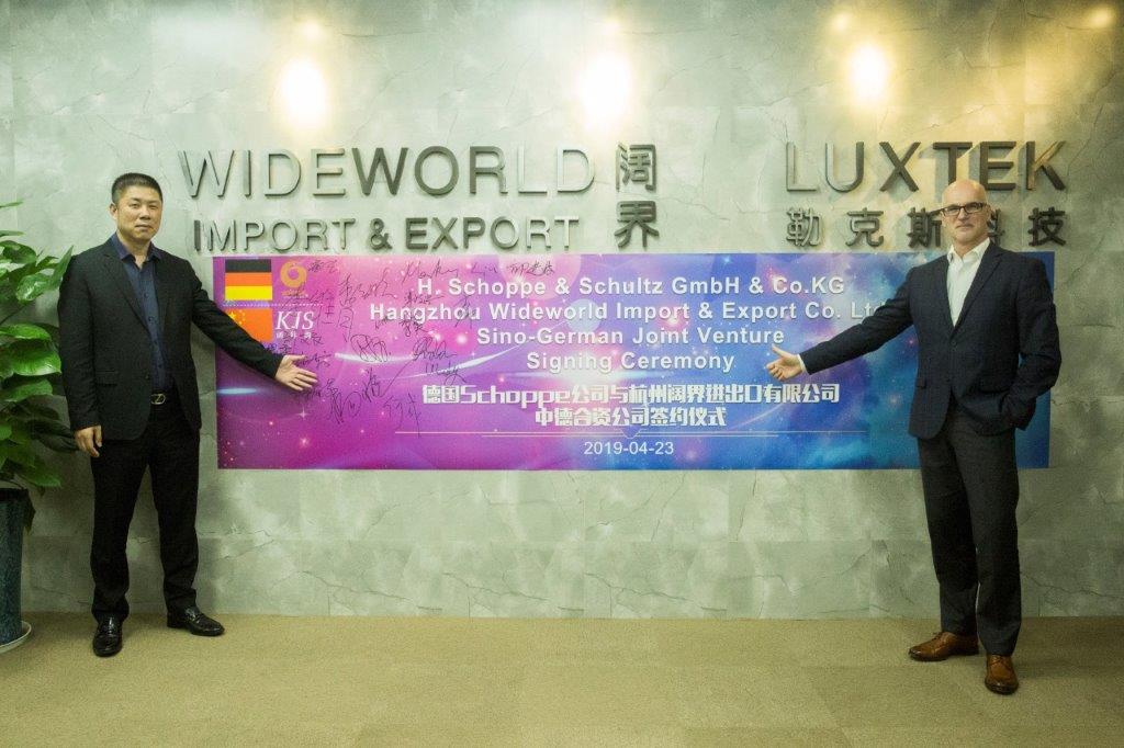 Two men stand beside a banner announcing a signing ceremony for a Sino-German joint venture between H. Schoppe & Schultz GmbH and Hangzhou Wideworld Import & Export Co. Ltd. The men are dressed in business attire. The setting appears to be a corporate office.