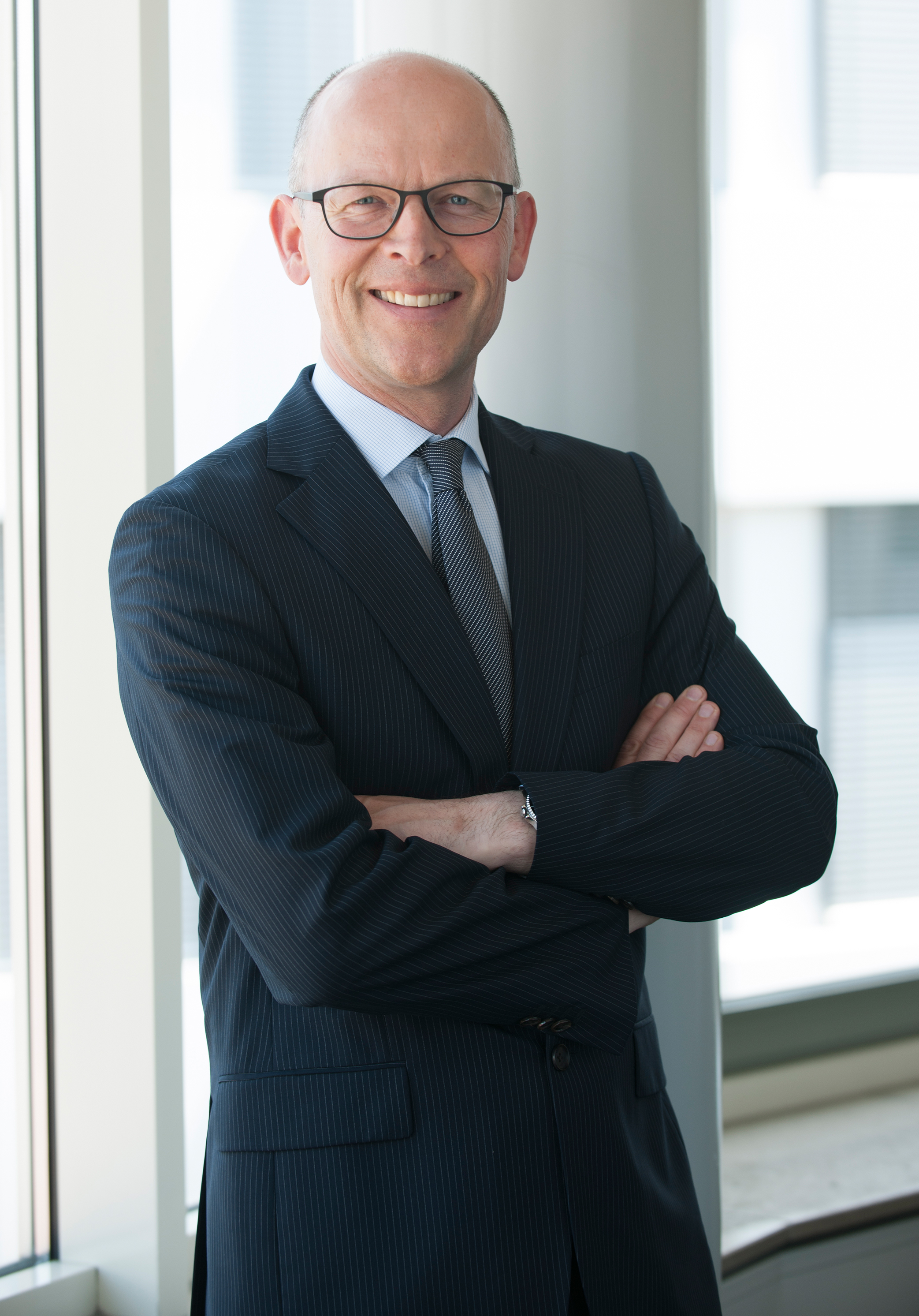 A man in a formal dark pinstriped suit stands confidently with arms crossed. He has short hair, glasses, and is smiling. The background features a bright, minimalist office environment with large windows.