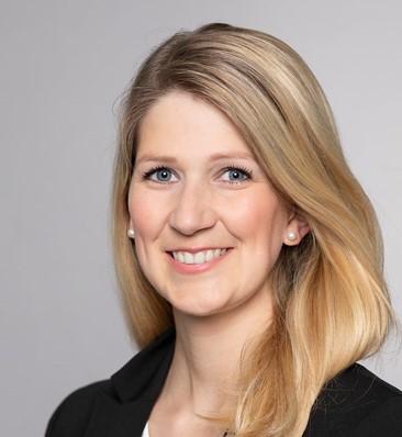 Porträt einer lächelnden Frau mit langen, blonden Haaren. Sie trägt einen schwarzen Blazer und hat hellblaue Augen. Der Hintergrund ist hellgrau, was den Fokus auf ihr freundliches Gesicht legt.