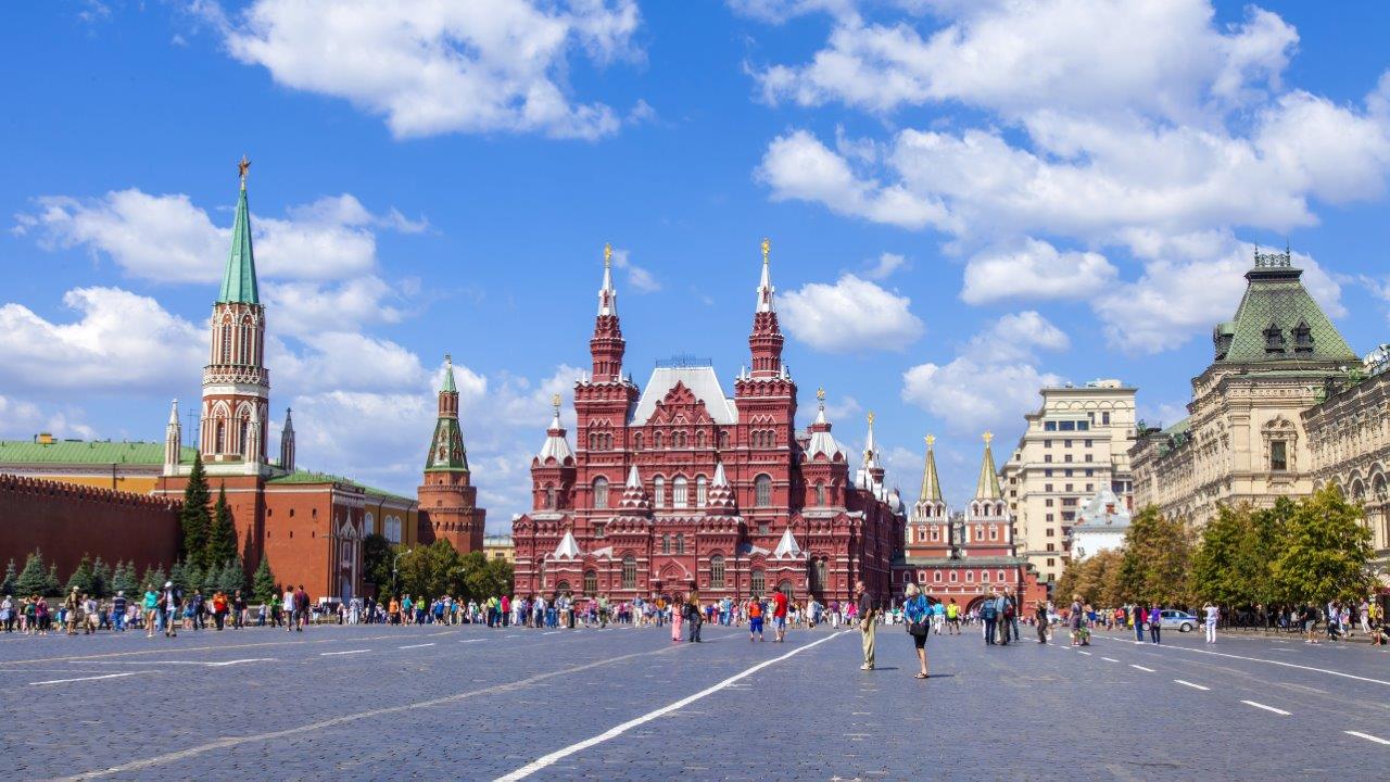 Auf dem Bild ist der Rote Platz in Moskau zu sehen, mit der beeindruckenden roten Backsteinarchitektur des historischen Museums im Vordergrund. Umgeben von Menschen, zeigt der Platz eine Vielzahl von Gebäuden, darunter die Zwiebeltürme des Kremls und moderne Bauwerke im Hintergrund. Der Himmel ist klar und blau.
