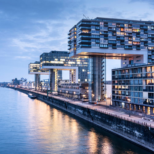 Blick auf moderne Architektur entlang einer Wasserstraße. Drei auffällige, schwebende Gebäude mit großen Fenstern reflektieren das Abendlicht. Der ruhige Wasserverlauf spiegelt die urbanen Strukturen wider, während die Umgebung eine Mischung aus städtischen und natürlichen Elementen zeigt.