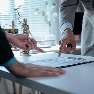 Zwei Personen stehen an einem Tisch und betrachten ein Dokument. Eine Hand zeigt auf den Text, während die andere Hand direkt neben dem Dokument liegt. Im Hintergrund sind unscharfe Gegenstände sichtbar, die auf ein Büroambiente hindeuten.