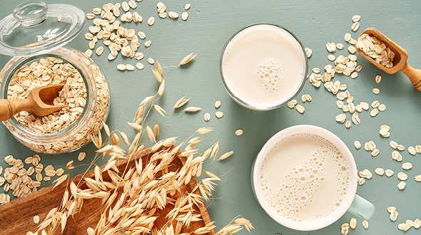 Eine Übersicht von Haferflocken, die auf einem hellblauen Tisch verteilt sind. Ein Glas mit Hafermilch und eine Tasse mit schaumiger Hafermilch sind sichtbar. Im Hintergrund steht ein Glasbehälter mit weiteren Haferflocken, umgeben von einigen ästhetisch angeordneten Haferhalmen.