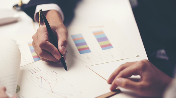 Ein Mann hält einen Stift und macht Notizen auf einem Blatt Papier mit Diagrammen und Grafiken. Eine zweite Hand ist ebenfalls sichtbar, möglicherweise beim Zeigen auf die Diagramme. Die Szene vermittelt einen geschäftlichen Kontext, wahrscheinlich eine Besprechung oder Analyse.