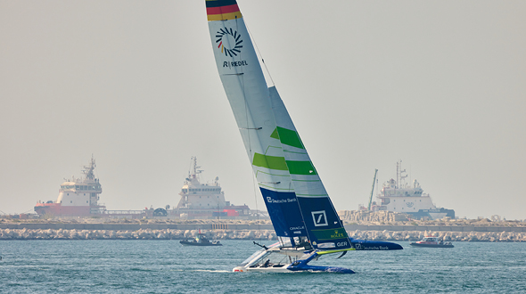 Ein modernes Segelboot mit einem großen, bunten Segel fährt über das Wasser. Im Hintergrund sind mehrere Schiffe und ein Felsen dezent angedeutet. Die Szene vermittelt ein Gefühl von Aktivität und maritimen Abenteuer.