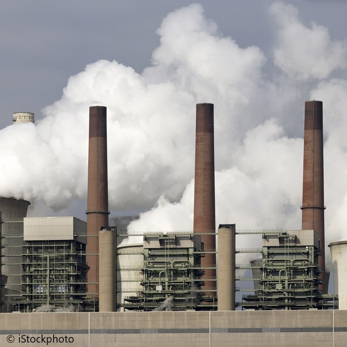 A power plant with multiple tall smokestacks emitting white steam and smoke into the sky. The industrial structure is partially obscured by clouds, emphasizing its scale and the environmental impact of its operations.