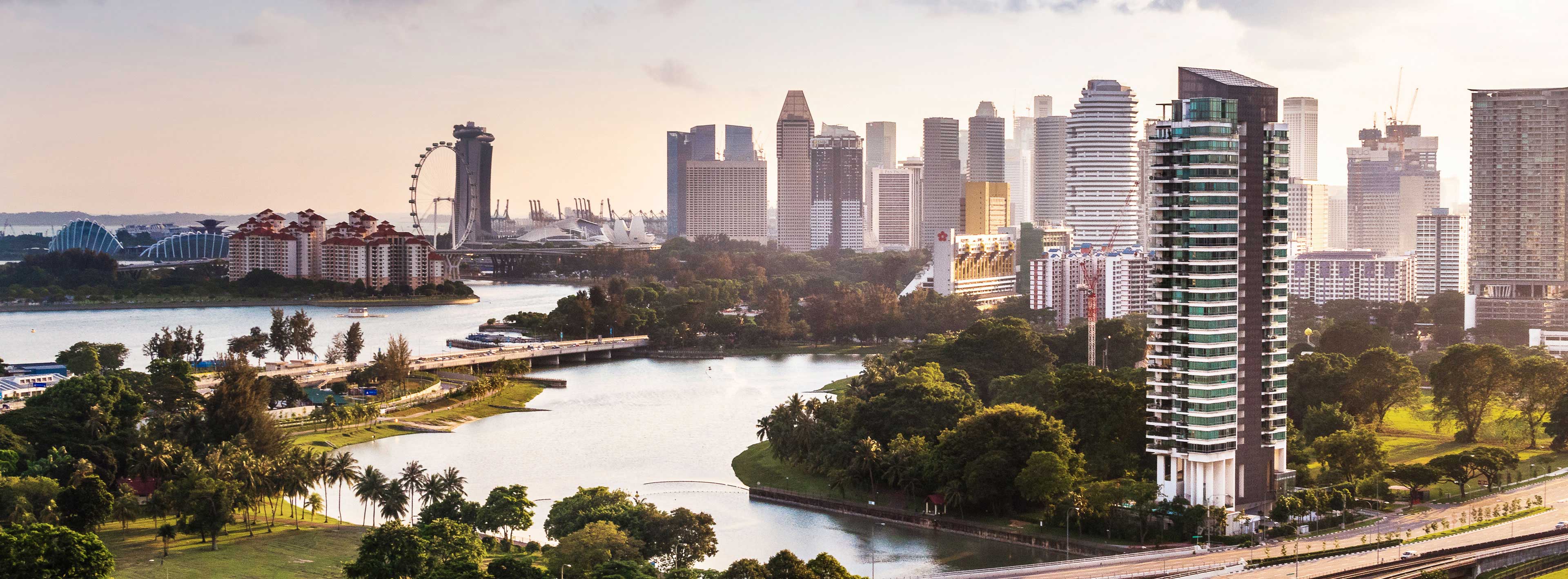 A panoramic view of a modern city skyline featuring tall buildings along a river. Lush greenery lines the waterfront, with a mix of urban architecture and nature. The scene is illuminated by soft, warm light, suggesting a late afternoon ambiance.