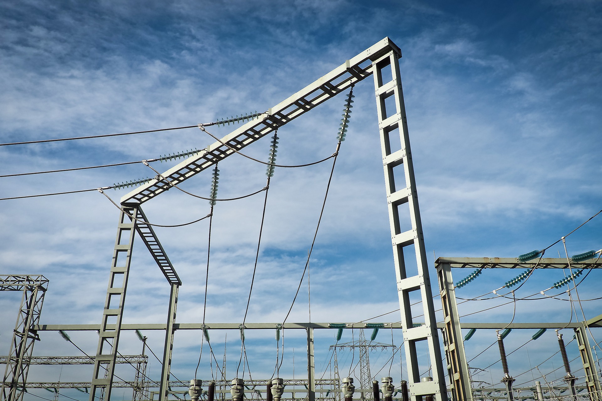 Eine elektrische Umspannstation ist zu sehen, mit hohen Metallstrukturen, die Stromleitungen halten. Über dem Gelände erstreckt sich ein blauer Himmel mit einigen Wolken. Die Anordnung der Kabel und Masten deutet auf eine industrielle Umgebung hin.