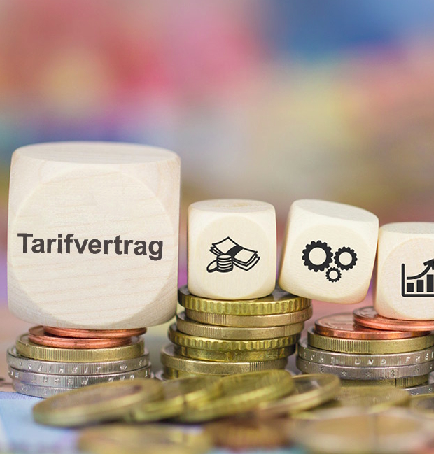 Holzwürfel mit Aufschrift "Tarifvertrag" und zwei weiteren Würfeln mit Symbolen für Geld, Zahnräder und ein Diagramm, angeordnet auf einem Stapel Münzen. Farbiger verschwommener Hintergrund.