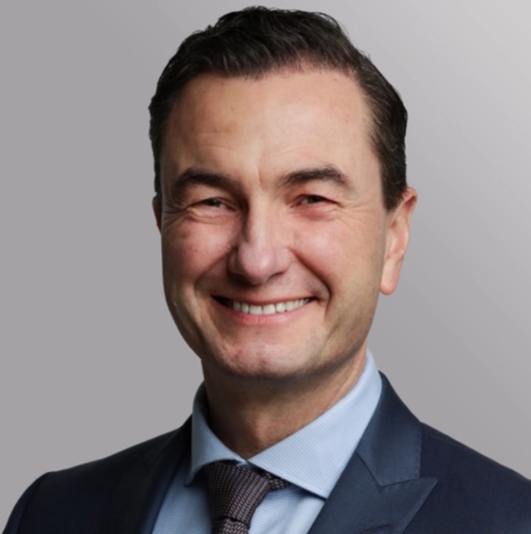 Ein lächelnder Mann in einem dunkelblauen Anzug mit einem hellen Hemd und einer gestreiften Krawatte. Sein kurzes, dunkles Haar ist glatt frisiert. Der Hintergrund ist grau.