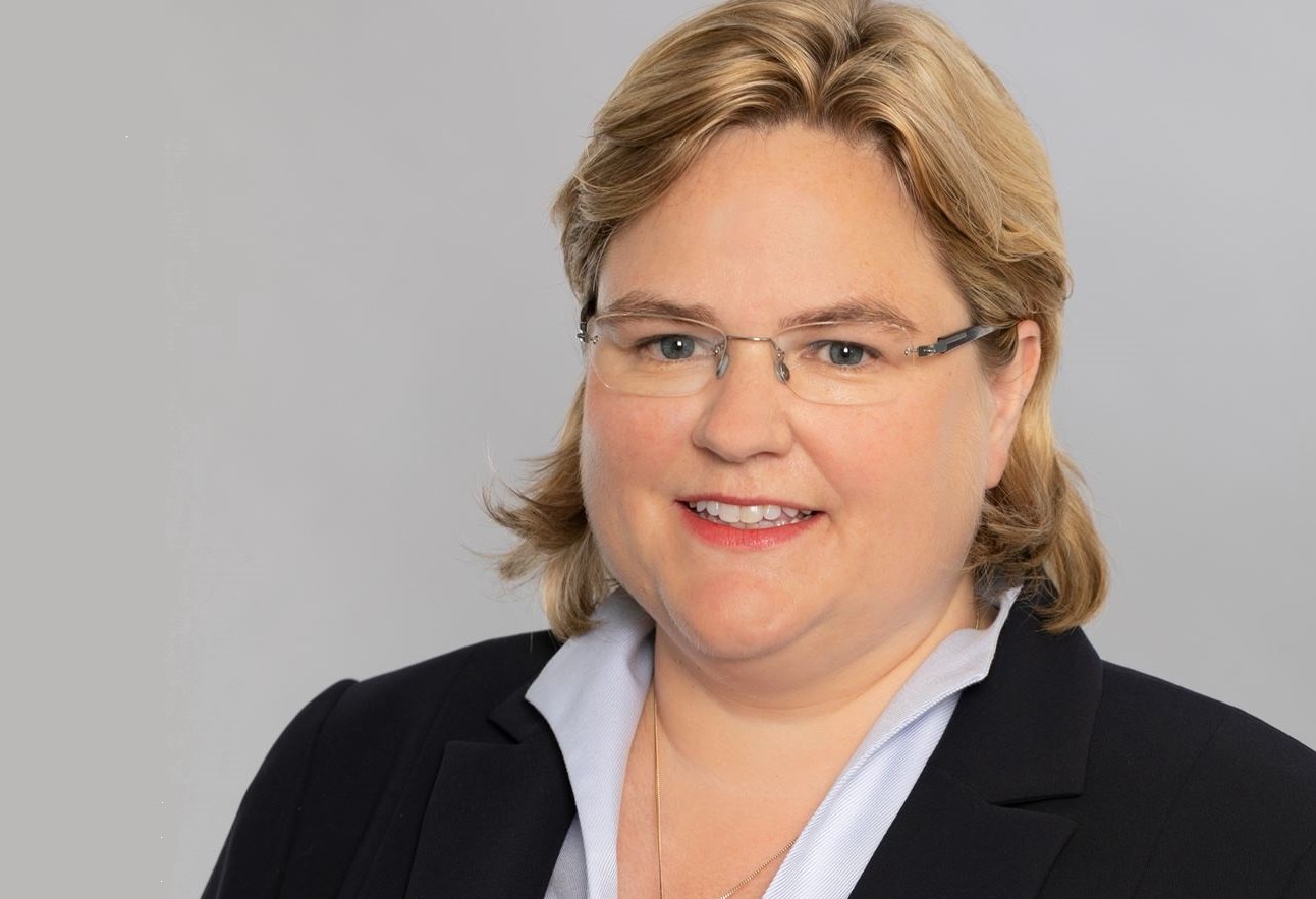 Ein Porträt einer Frau mit blonden, schulterlangen Haaren, die einen schwarzen Blazer und ein hellblaues Hemd trägt. Sie lächelt freundlich und trägt eine kleine Brille. Der Hintergrund ist einheitlich grau.
