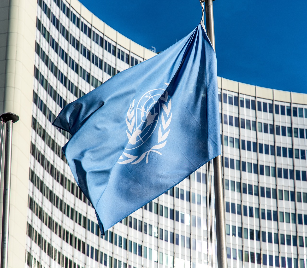 Die Fahne der Vereinten Nationen weht vor einem modernen Gebäude. Die Flagge ist himmelblau mit dem Symbol der UN, das eine Weltkarte in einem olivenblattumrahmenden Ring zeigt. Der Himmel ist klar und der Wind bewegt die Fahne leicht.
