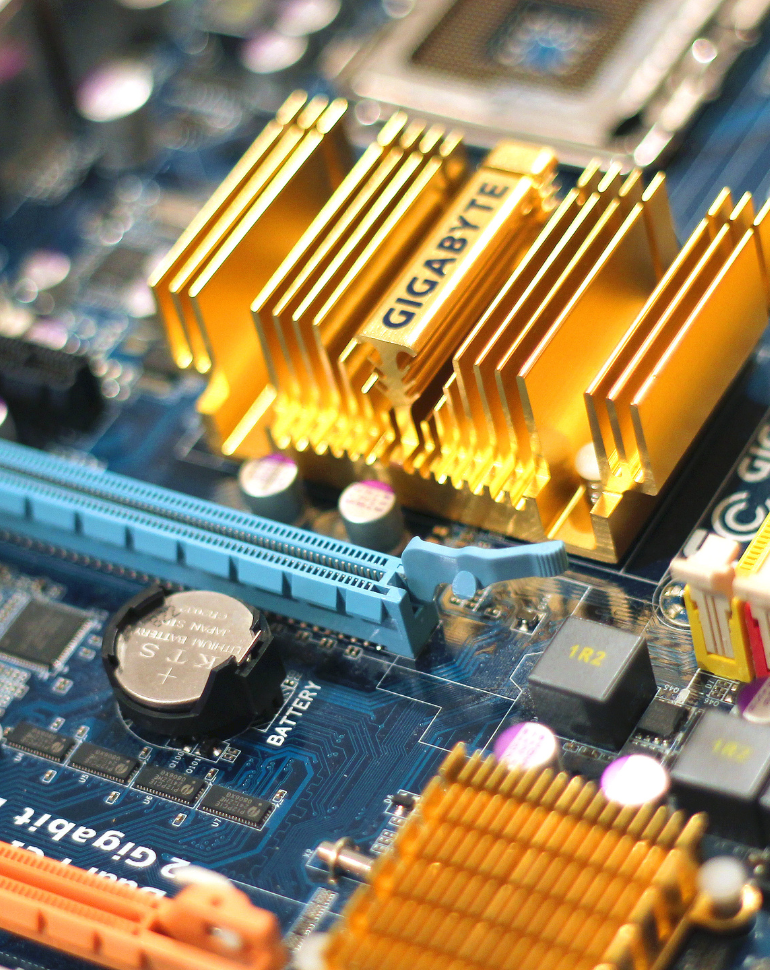 Nahaufnahme eines Motherboards mit goldenen Kühlkörpern, die das Logo "GIGABYTE" tragen. Zu sehen sind verschiedene elektronische Komponenten, darunter ein Speichersteckplatz und ein Batterieanschluss. Die Leiterbahnen sind auf einem blauen Hintergrund angeordnet.