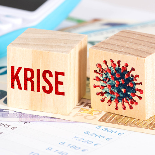 Zwei Holzklötze sind abgebildet. Auf dem linken steht in roter Schrift das Wort "KRISE". Der rechte block zeigt eine grafische Darstellung des Coronavirus mit roten Stachelstrukturen. Hintergrund ist ein Haufen von Geldscheinen.