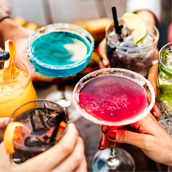 Sechs Hände halten verschiedene Cocktails, darunter ein blauer Drink, ein roter Drink mit Zuckerrand, ein grüner Drink und ein dunkler Drink. Die Getränke sind bunt und scheinen in einer geselligen Umgebung serviert zu werden, was eine festliche Atmosphäre vermittelt.