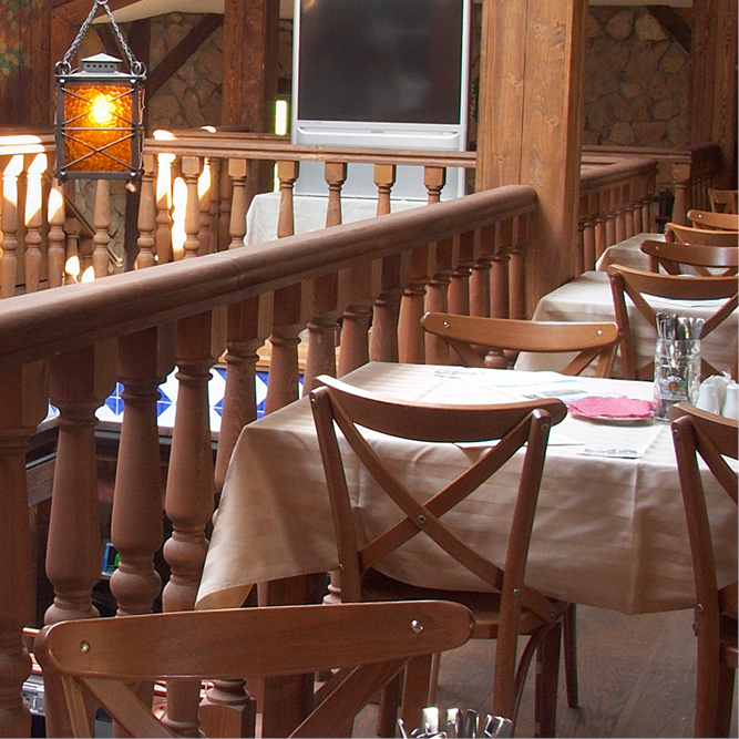 A cozy restaurant interior featuring wooden railings and tables set with light tablecloths. The ambiance is warm with soft lighting from an overhead lantern. The layout suggests a welcoming atmosphere for dining, enhanced by rustic wooden accents and a hint of decorative stonework in the background.