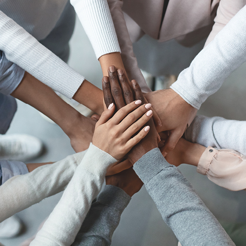 A diverse group of hands of varying skin tones is stacked together in a circular arrangement, symbolizing unity and collaboration. The hands display different styles and tones, reflecting inclusivity and teamwork.