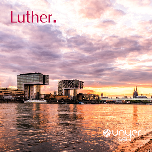 Der Blick auf den Fluss zeigt moderne Gebäude am Ufer, während der Himmel in warmen Farben zum Sonnenuntergang leuchtet. Im Hintergrund sind kirchliche Türme zu erkennen. Der Fokus liegt auf der harmonischen Verbindung von Architektur und Natur.