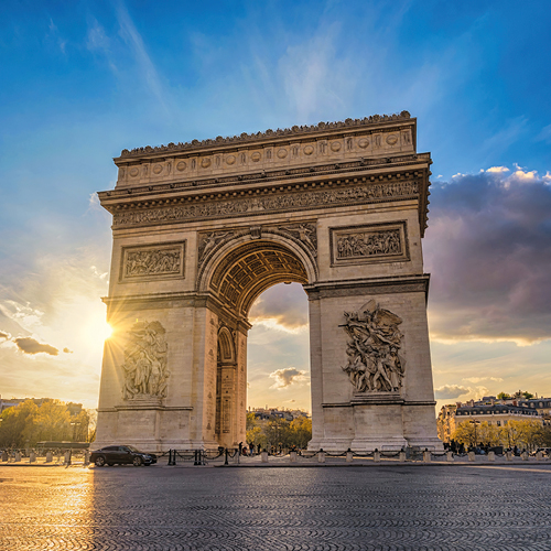 The image features the Arc de Triomphe in Paris, illuminated by the setting sun. The monumental arch displays intricate sculptures on its sides, standing majestically against a backdrop of blue and golden skies. The scene captures both historic architecture and a beautiful sunset.