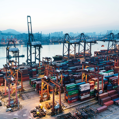 Ein Blick auf einen großen Hafen mit zahlreichen Containern und Kränen. Die Container sind in verschiedenen Farben gestapelt, während Kräne bereitstehen, um den Warenumschlag zu unterstützen. Im Hintergrund ist das Wasser des Hafens und eine Stadtlandschaft sichtbar.