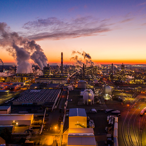 Eine Luftaufnahme einer Industrieanlage bei Sonnenuntergang. Rauch und Dampf steigen aus hohen Schornsteinen auf, während die Gebäude beleuchtet sind. Der Himmel zeigt warme Farben, die Kontraste zwischen Natur und Industrie betonen.